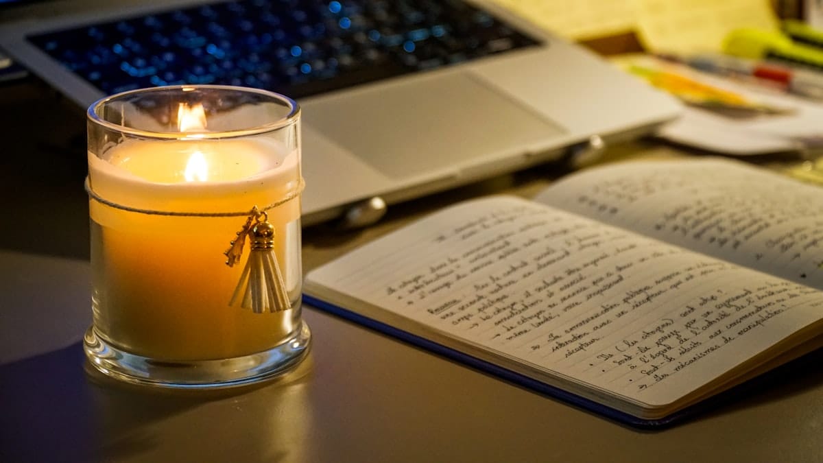 A lit candle beside an open notebook on a desk in the evening
